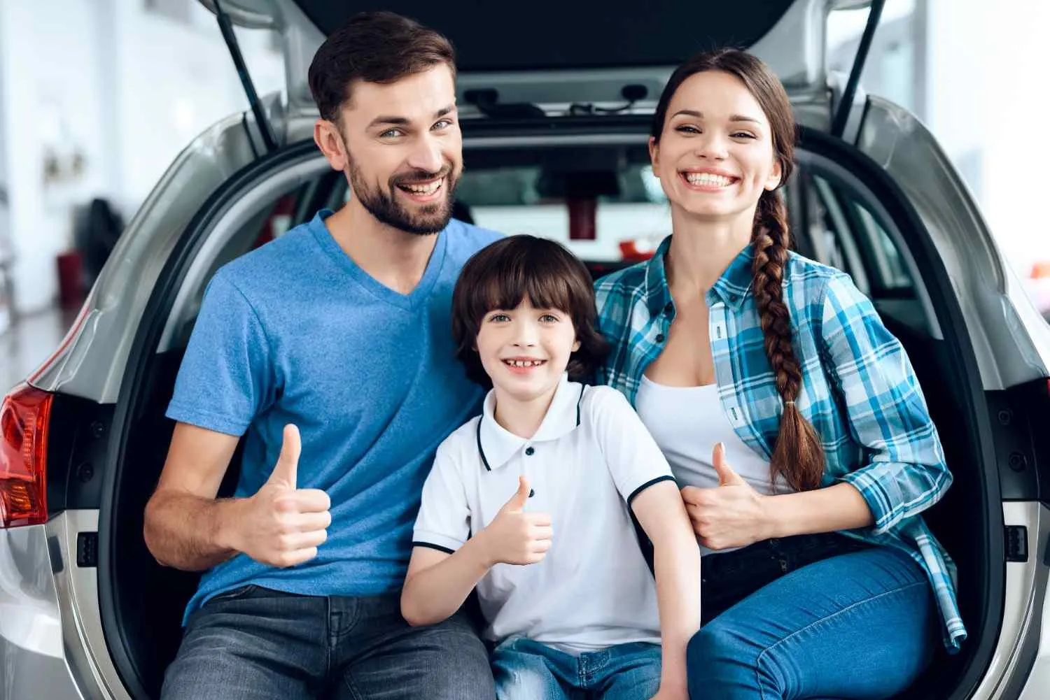 A family of three sitting in the open back of their car, all giving a thumbs up